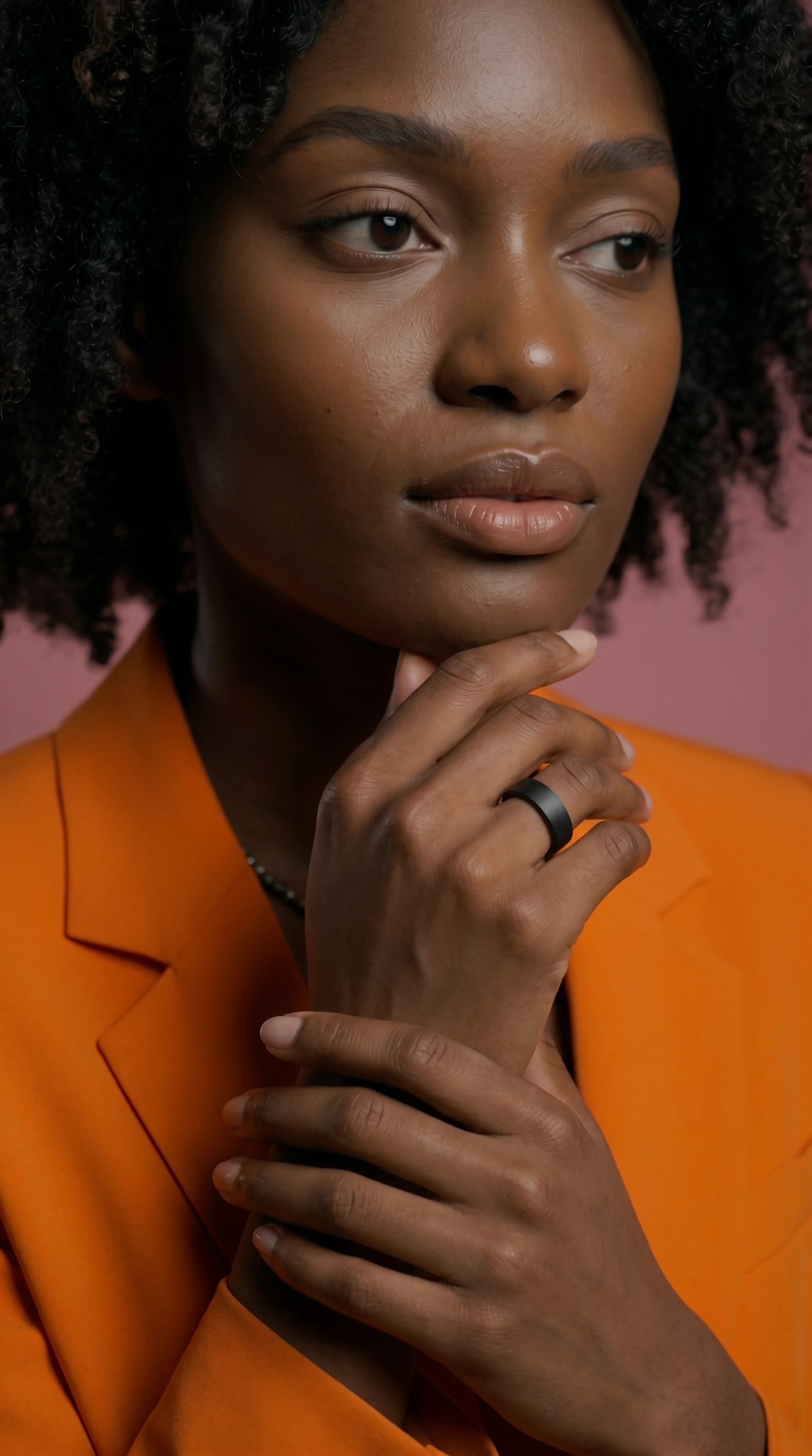 Editorial-Closeup: Frau mit MYnextRING am Finger, Hand am Kinn, oranger Blazer auf rosa Studio-Hintergrund